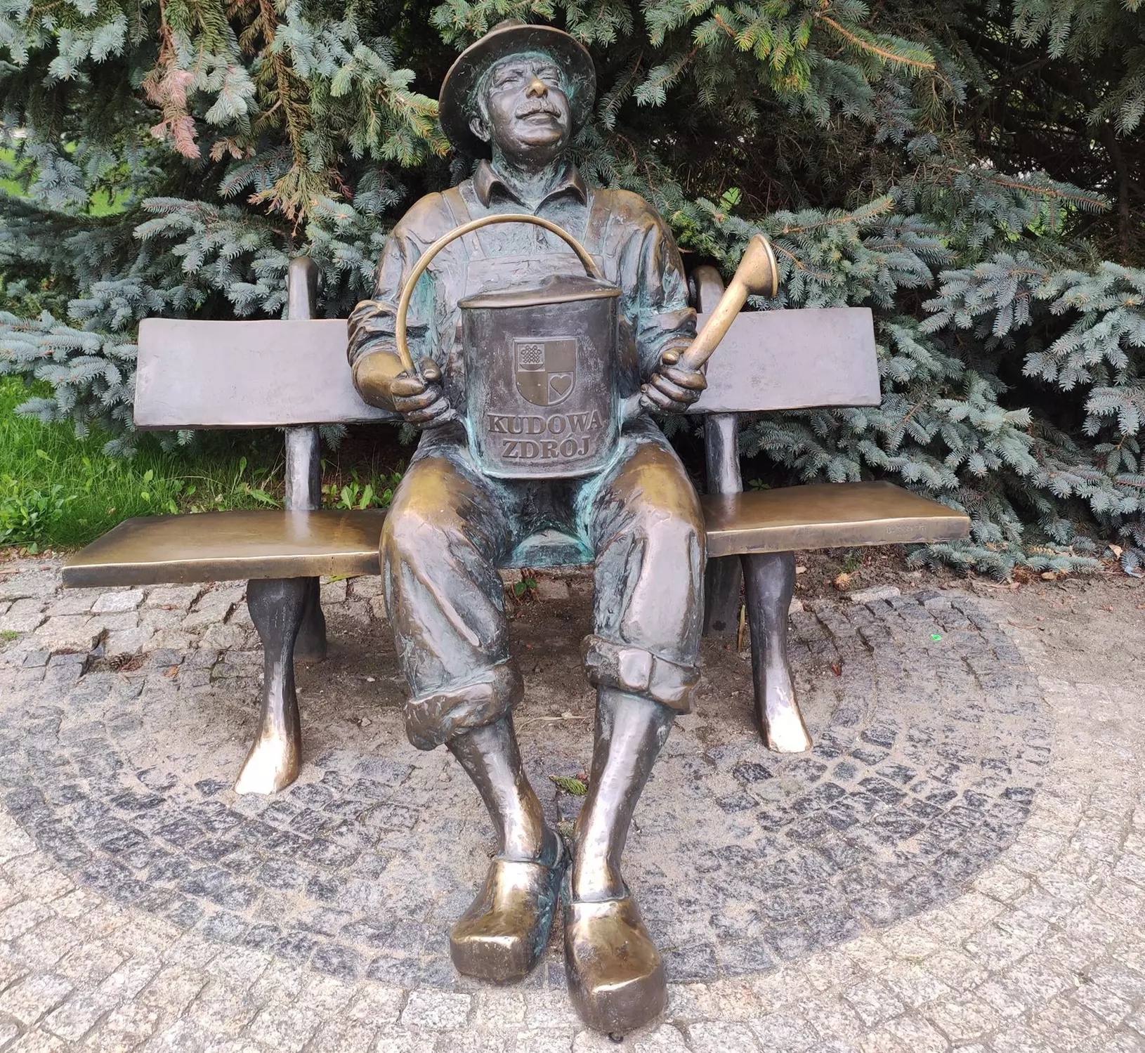 Na jiné bronzové lavičce v Kudowě-Zdróji sedí zahradník s konví, na níž je městský erb